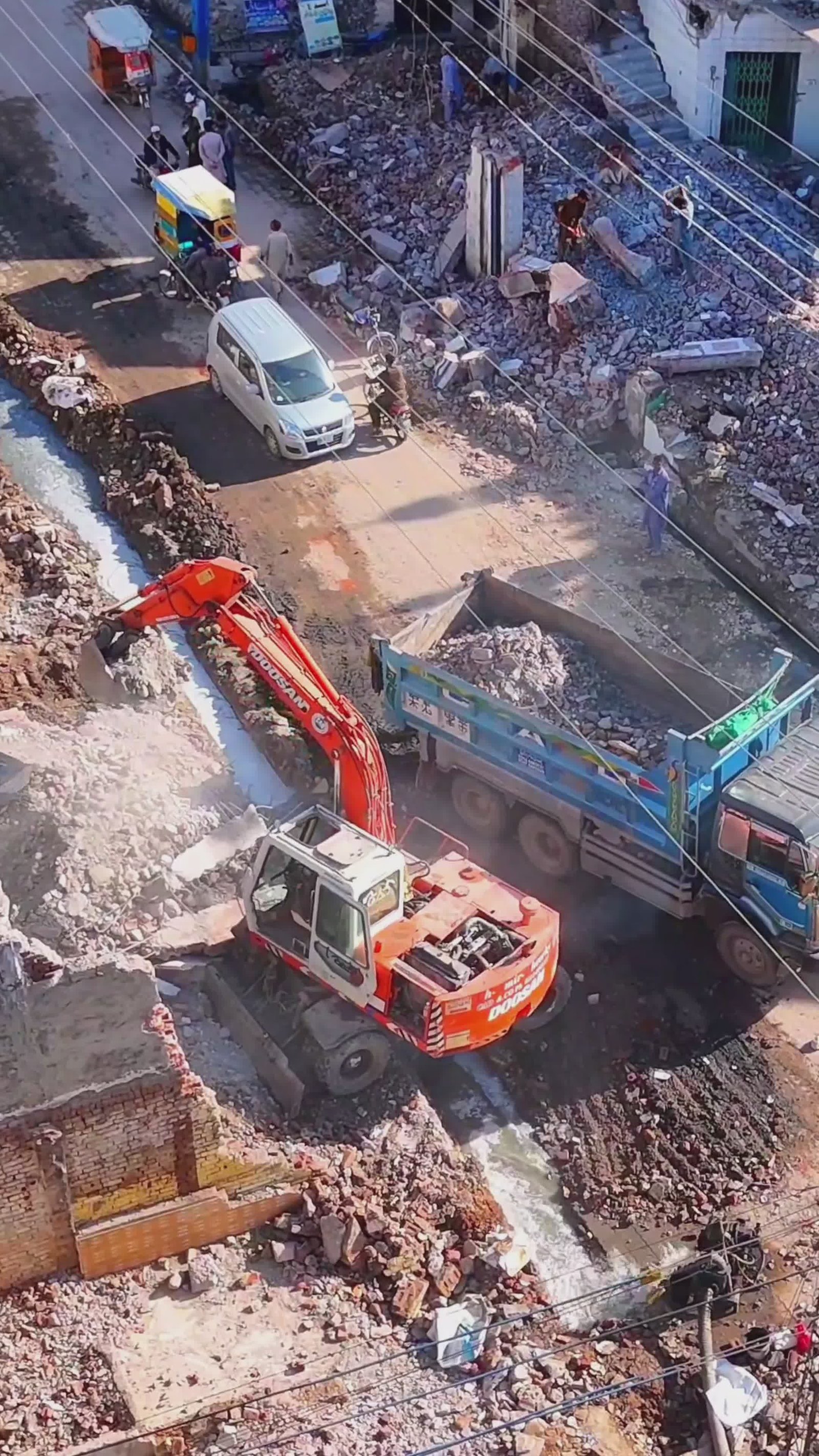 Chantier de construction avec excavatrice orange déversant des gravats dans une benne; rue encombrée et câbles électriques.