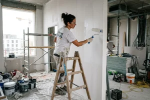 Femme peintre en bâtiment sur une échelle, appliquant de la peinture sur un mur dans un chantier, matériel et seaux autour. Fiche métier peintre en bâtiment.