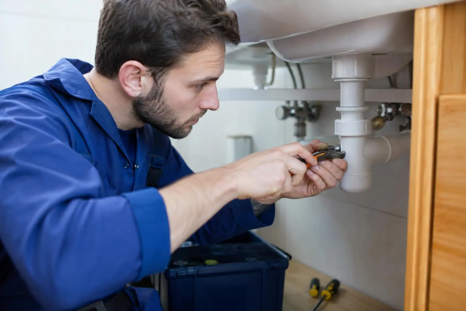 Homme en bleu travaillant sous l’évier, serrant un raccord de plomberie pour réparer une fuite, sous un meuble de cuisine. fiche métier plombier intégrée.